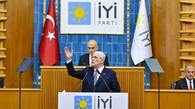 Dervişoğlu kürsüde elini kaldırmış şekilde konuşuyor, önünde İYİ yazıyor. Arkasında ve yanında birer kişi oturmuş durumda.