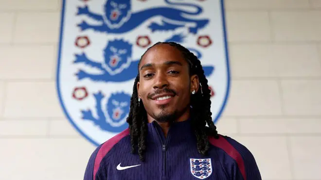 Djed Spence smiling in front of a giant England badge.