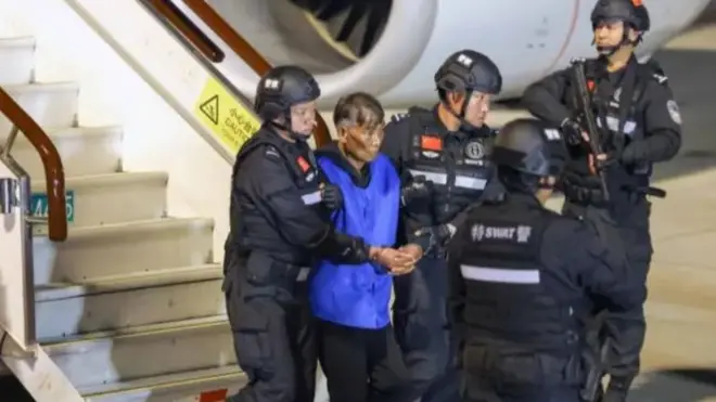 Một người đàn ông lớn tuổi mặc áo vest màu xanh đang bị cảnh sát dẫn giải ra khỏi máy bay