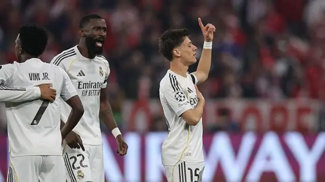 Arda güler ayakta, gözleri kapalı, sol elini kalbine koymuş, sağ elinin işaret parmağı gökyüzünü gösteriyor.