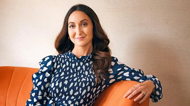 Eve Simmons é uma mulher branca, de cabelos longos e castanhos. Ela usa um vestido azul e está sentada em um sofá laranja.