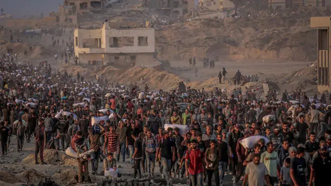 大批人群,其中一些人扛著麻袋,走過加沙被毀的建築物