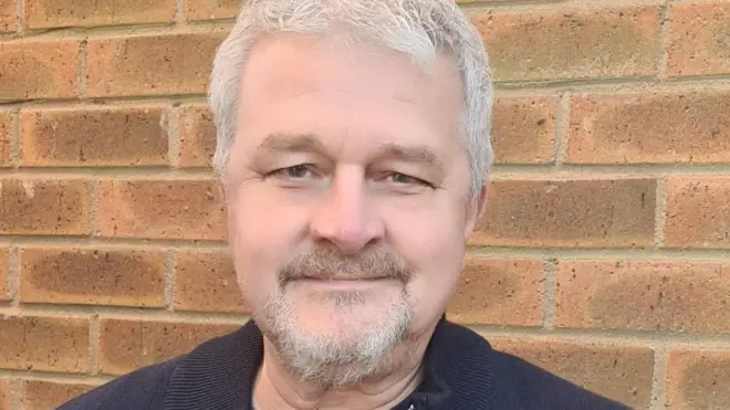 David, un hombre con cabello gris y una pequeña barba gris, que viste una camiseta negra, mira a la cámara.