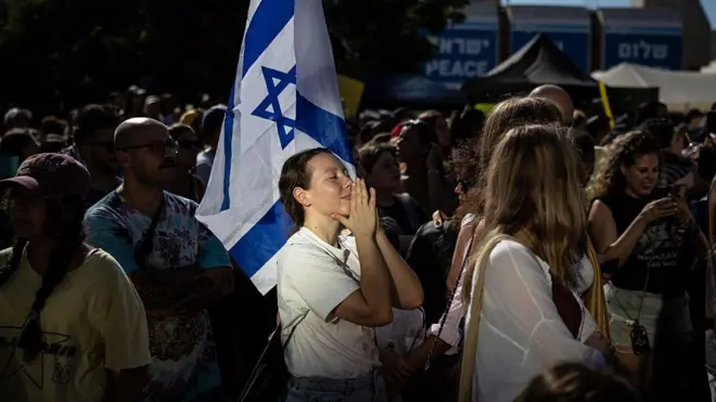 Bir kadın ellerini dua eder gibi kapamış, gözleri de kapalı, etrafından çok sayıda kişi var.