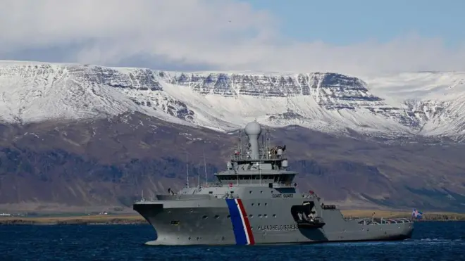 Uma embarcação da guarda costeira islandesa na água, com uma paisagem de montanhas nevadas ao fundo.