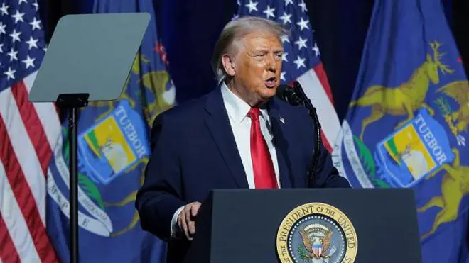 Trumo bir kürsüden konuşma yapıyor, arka planda ABD bayrağı var. 