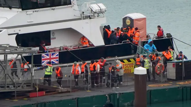 オレンジ色の救命胴衣を着た20人以上の人たちが、イギリス国旗がついた船から降ろされ、埠頭（ふとう）に立っている