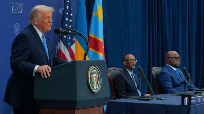 Perezida w'Amerika Donald Trump ahagaze arimo kuvuga ijambo. Yambaye ikositimu y'ubururu bwijimye, ishati y'umweru na karuvati y'ubururu. Iruhande rwe hari ibendera ry'Amerika, iry'u Rwanda n'irya Repubulika ya Demokarasi ya Congo. Hirya imbere y'ameza hicaye Perezida w'u Rwanda Paul Kagame na Perezida wa DRC Félix Tshisekedi bambaye kositimu n'indorerwamo, bareba imbere, bijimye mu maso. Ifoto yo ku wa kane, ku itariki ya 4 Ukuboza (12) mu 2025, i Washington DC muri Amerika.