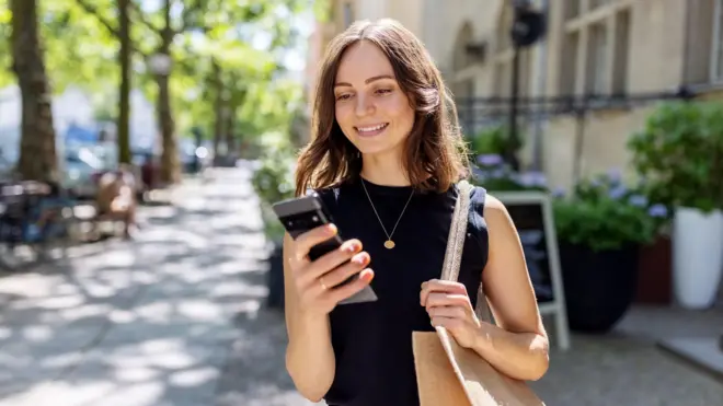 Młoda kobieta uśmiecha się, patrząc w telefon. Jest na zewnątrz, a w tle zdjęcia widać drzewa.