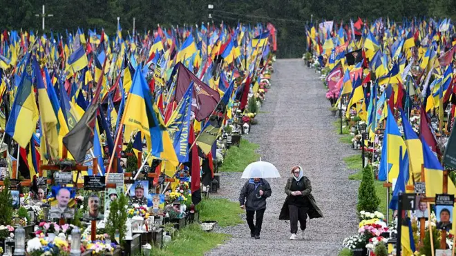 Під дощем двоє людей ідуть алеєю між могилами українських військових, прикрашених численними синьо-жовтими та червоно-чорними прапорами, квітами й портретами полеглих.