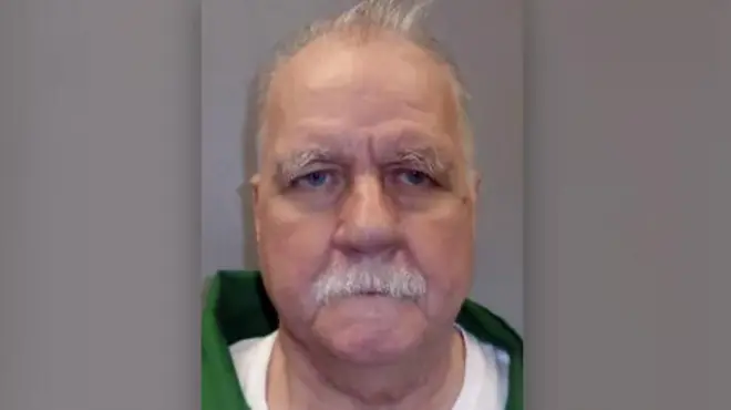 The prisoner is seen in a green jacket with a white shirt. He has a white moustache and white hair and there is a grey background