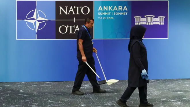NATO yazan mavi panonun önünde iki temizlik görevlisi