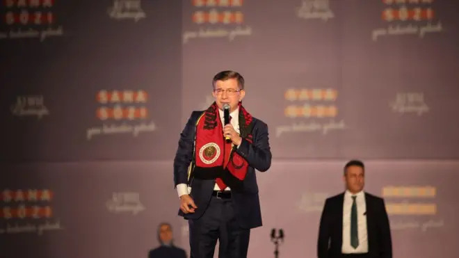Davutoğlu kırmızı bir atkıyla bir miting sahesinde elinde mikrofonlar ayakta, resmi giyimli. 