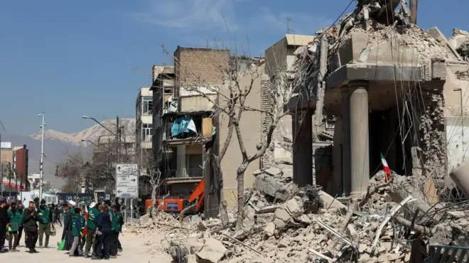 激しく損壊した建物があり、その前の道路に人々が集まっている。青空が広がり、遠くには雪山の山並みが見える