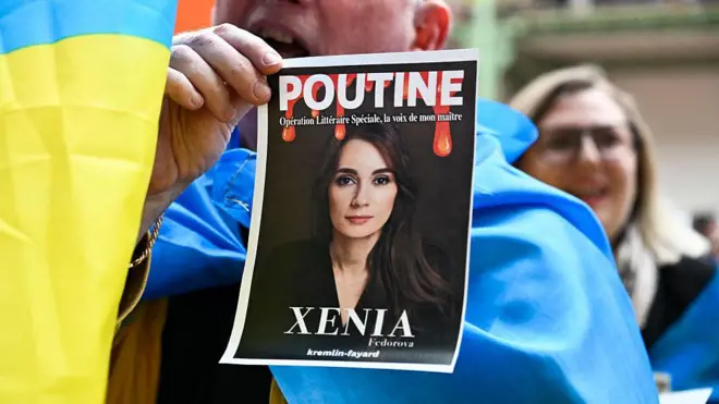 Сторонники Украины устроили акцию протеста на презентации книги Ксении Федоровой в прошлом апреле в Париже - участник акции держит фото Федоровой с надписями "Путин" и "Ксения"