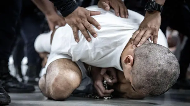 Erkek bir mahkûm yerde otururken, yukarıdan gelen eller vücudunu ve başını yere doğru bastırıyor. Başı kazınmış olan mahkûm, beyaz bir tişört ve gri şort giyiyor; elleri ise kelepçeli.