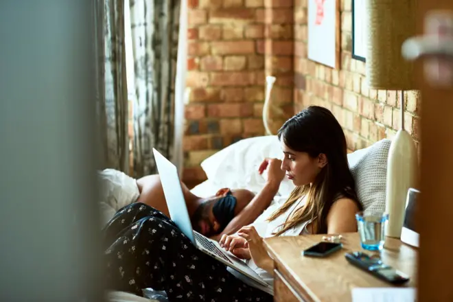 امرأة شابة تستخدم الكمبيوتر المحمول في السرير بينما ينام شريكها