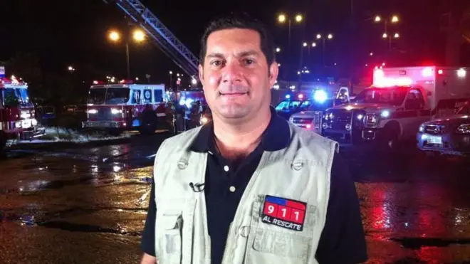 Foto mostra um homem branco com jaqueta em frente a caminhões dos bombeiros