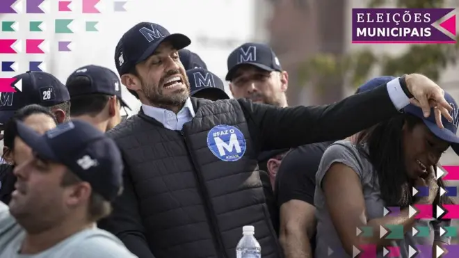 A foto mostra Pablo Marçal durante a campanha, vestindo um colete escuro e boné com a letra M, rodeadoapostas esportivas pdfpessoas com o mesmo boné.