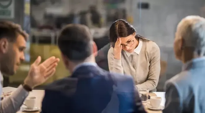 Mulher branca segura a cabeça e tem cara de frustração em frente a homens brancos em local de trabalho