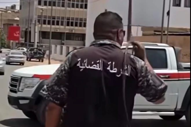 صورة من التسجيل المصور تُظهر أحد عناصر الشرطة القضائية واقفاً من الخلف، مرتدياً زياً شرطياً يحمل عبارة "الشرطة القضائية"، وممسكاً سوطاً بيده اليمنى قبل البدء في جلد المرأة.