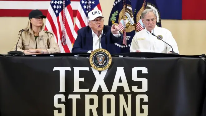 星条旗とテキサス州旗を背にした3人が、「TEXAS STRONG（強いテキサス）」と書かれた幕で覆われたテーブルに向かって座っている。