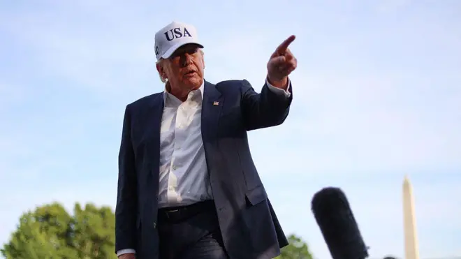الرئيس الأمريكي دونالد ترامب يشير بيده أثناء حديثه مع صحفيين بعد وصوله إلى الحديقة الجنوبية للبيت الأبيض في واشنطن العاصمة. يرتدي بدلة رسمية كحلية اللون وقميصاً أبيض دون ربطة عنق، وقبعة بيضاء كتب عليها "الولايات المتحدة الأمريكية" - USA باللغة الإنجليزية باللون الأسود.