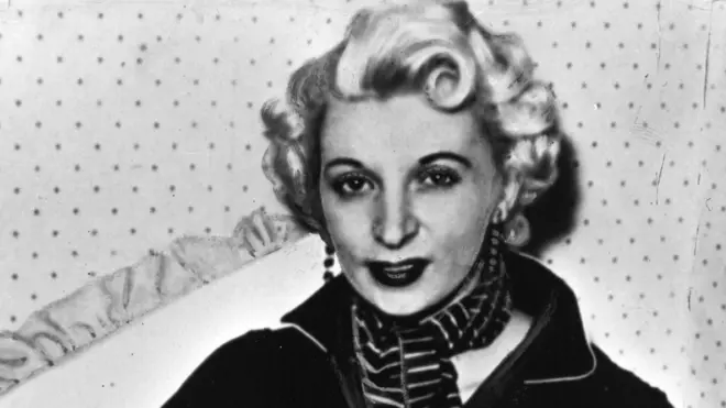 Retrato de Ellis, rubia, pelo corto ensortijado, maquillada, con aretes largos, un pañuelo en la garganta y un sweater oscuro de cuello alto abierto. Recostada en un cojín, contra una pared con papel de colgadura de puntos.