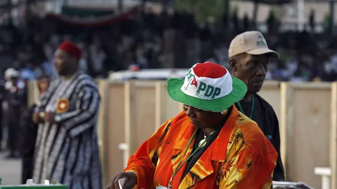 Woman dey slot in paper into ballot, she wear hat wey get PDP ontop