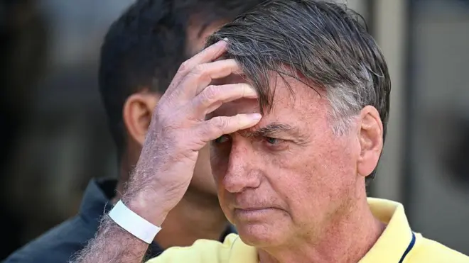 Jair Bolsonaro mirando hacia la izquierda y con la mano derecha sobre la frente. Viste una camiseta con cuello amarilla.