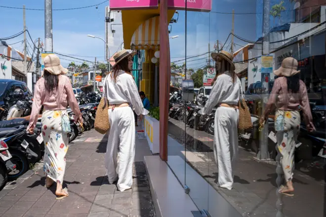 Dwie młode kobiety w letnich strojach i kapeluszach idą ulicą. Jedna z nich patrzy w witrynę sklepową. Na krawężniku rzędami zaparkowano skutery i samochody. Kuta, Bali, Indonezja. 5 marca 2026 r. 