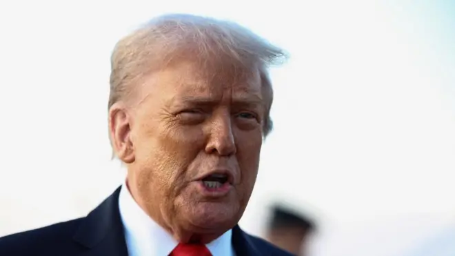 Donald Trump, wambaye ikositimu yijimye na karuvati itukura, arimo kuvuga areba muri 'camera'. 