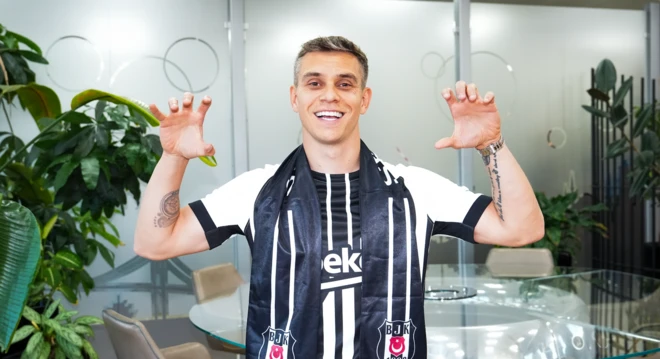 Siyah beyaz Beşiktaş atkılı bir futbolcu gülere poz veriyor. Ellerini kartal tutuşu şeklinde iki yana açmış