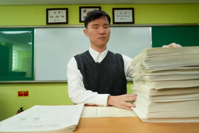 서울맹학교 한동현 군이 점자로 된 모의고사 시험지를 손으로 살펴보고 있다. 높게 쌓여있는 점자 시험지 옆에는 일반 지필용 시험지가 있다.