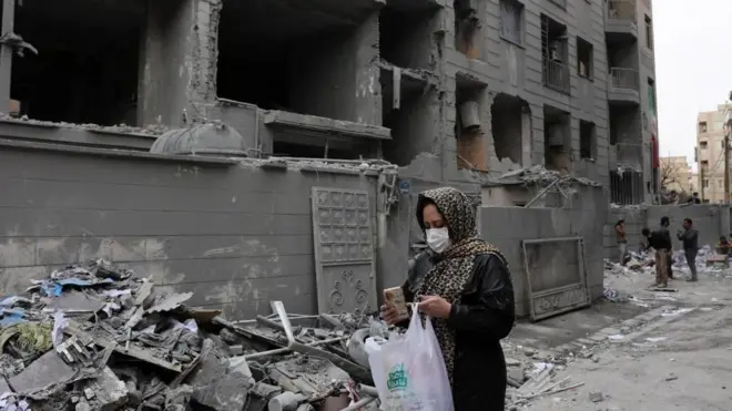 がれきの前を、マスクをした女性がスマートホンを見ながら歩いている。手には白いプラスチックバッグを持っている