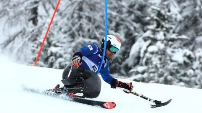 白いヘルメット、ゴーグル、青いウエアを着け、チェアスキーに座った鈴木が、先端に短いスキーがついたアウトリガーを両手に持ちながら滑っている