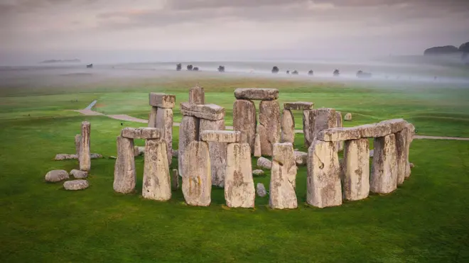 Resim Stonehenge'i gösteriyor. Arkasında antik anıtın tam dairelerinin ön planda olduğu sisli bir gökyüzü var. Dış dairenin bir parçasını oluşturan, her biri üstte başka bir büyük taş bulunan iki büyük dik taştan oluşan dört set. İç alanın daha büyük taşları dış çemberinkilerin üzerinde görülebilir. Yeşil çim anıtı her taraftan çevreliyor.  