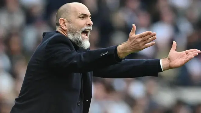 Koyu renk takım elbise giymiş sakallı Igor Tudor ellerini iki yana açmış halde bağırıyor, arka planda bulanık halde staddaki seyirciler görülüyor