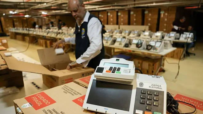 urna eletrônica sobre uma caixa. No fundo, várias urnas e um funcionário da Justiça Eleitoral. 