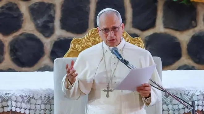  白いカソック（祭服）姿で胸に銀色の十字架をかけたレオ14世が、教会の祭壇を背に、紙の原稿を手に、マイクを前に、聴衆に向かって話している