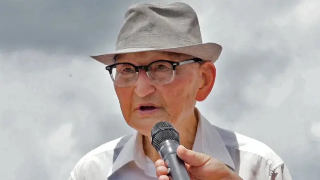 Ahn Hak-sop, wearing glasses and a hat, speaks with a mic being held close to his mouth