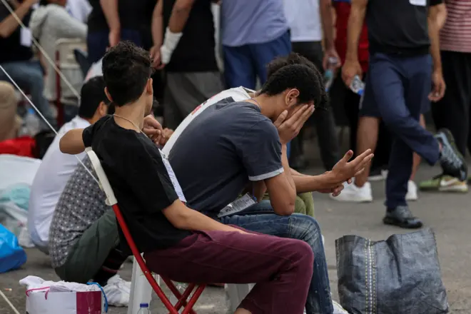 Homens em área portuária, alguns sentados, outros em pé - e um deles com aos mãos no rosto, expressando preocupação