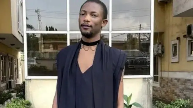 Ezra wear black robe and stand in front of one building surrounded by flowers. E wear choker round im neck and also wear lipstick.