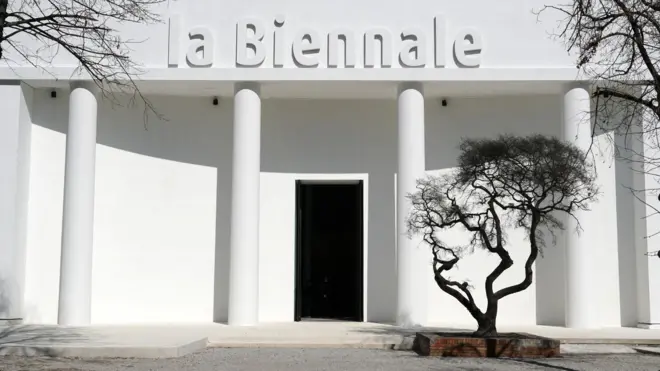 Bela zgrada sa natpisom „la Biennale“ i crnim drvetom ispred nje
