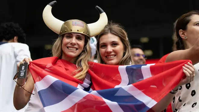 Norveç'in kırmızı mavi beyaz renklerinde, haç şeklindeki bayrağını gülerek tutan iki kadın taraftar. Birinin kafasında ülkenin sembolü Vikinglerin şapkası var.