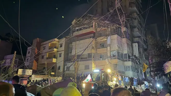 一部が崩壊した建物の近くで夜間、人々や重機が照明に照らされている。それを離れたところから多くの人々が見ている