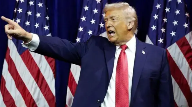 Aarẹ Donald Trump, o n sọrọ lọwọ, o naka rẹ kan siwaju ninu aṣọ oyinbo alawọ aro, itẹnu funfun ati tai pupa to de sọrun. Asia America wa lẹyin rẹ.