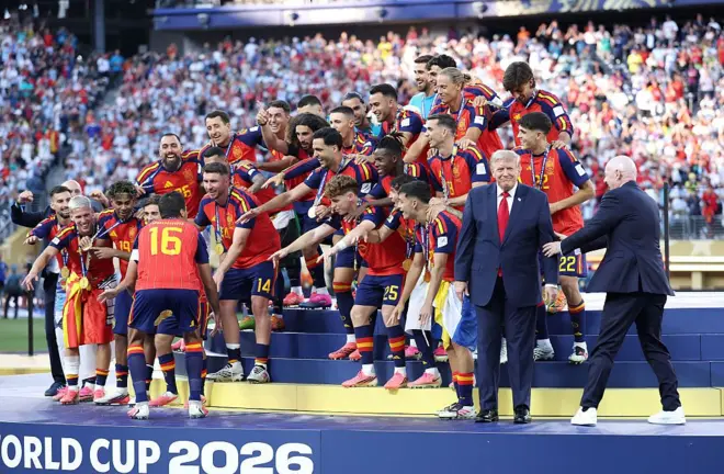 رودري، لاعب المنتخب الإسباني رقم 16، يستعد لرفع كأس العالم لكرة القدم، بينما يوجه رئيس الفيفا جياني إنفانتينو الرئيس الأمريكي دونالد ترامب، للخروج من أمام المنتخب الإسباني المحتفل بفوزه.
يرتدي ترامنب وانفانتينو بدلات رسمية، بينما يرتدي لاعبو المنتخب الإسباني لباس منتخب بلادهم الرياضي بالألوان الأحمر والكحلي والأصفر، يحتفلون وخلفهم الجماهير.