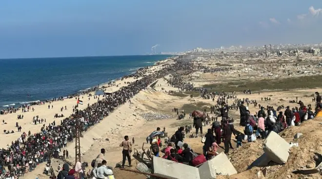 آلاف النازحين يعودون إلى شمال غزة بعد وقف إطلاق النار، في الصورة الملتقة نهاراً الناس بأعداد كبيرة يقفون قرب الشاطئ، ويرفع أحدهم العلم الفلسطيني على برج يبدو أنه للاتصالات أو الكهرباء