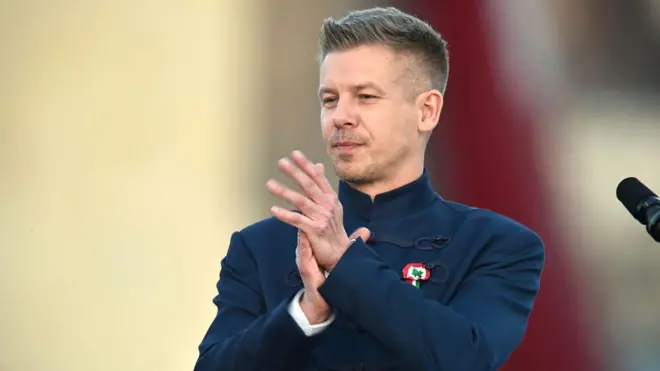 Lacıvert takım giyen bir sarışın erkek ellerini kavuşturmuş. Alkışlıyor gibi görünüyor.